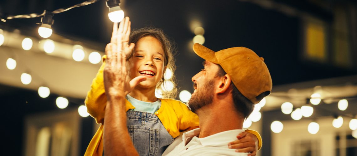 Proud Handsome Father Helping His Little Beautiful Daughter to Change a Lightbulb in Fairy Lights Backyard Installation at Home. Father and Daughter High Five and Celebrate Successful Fix.